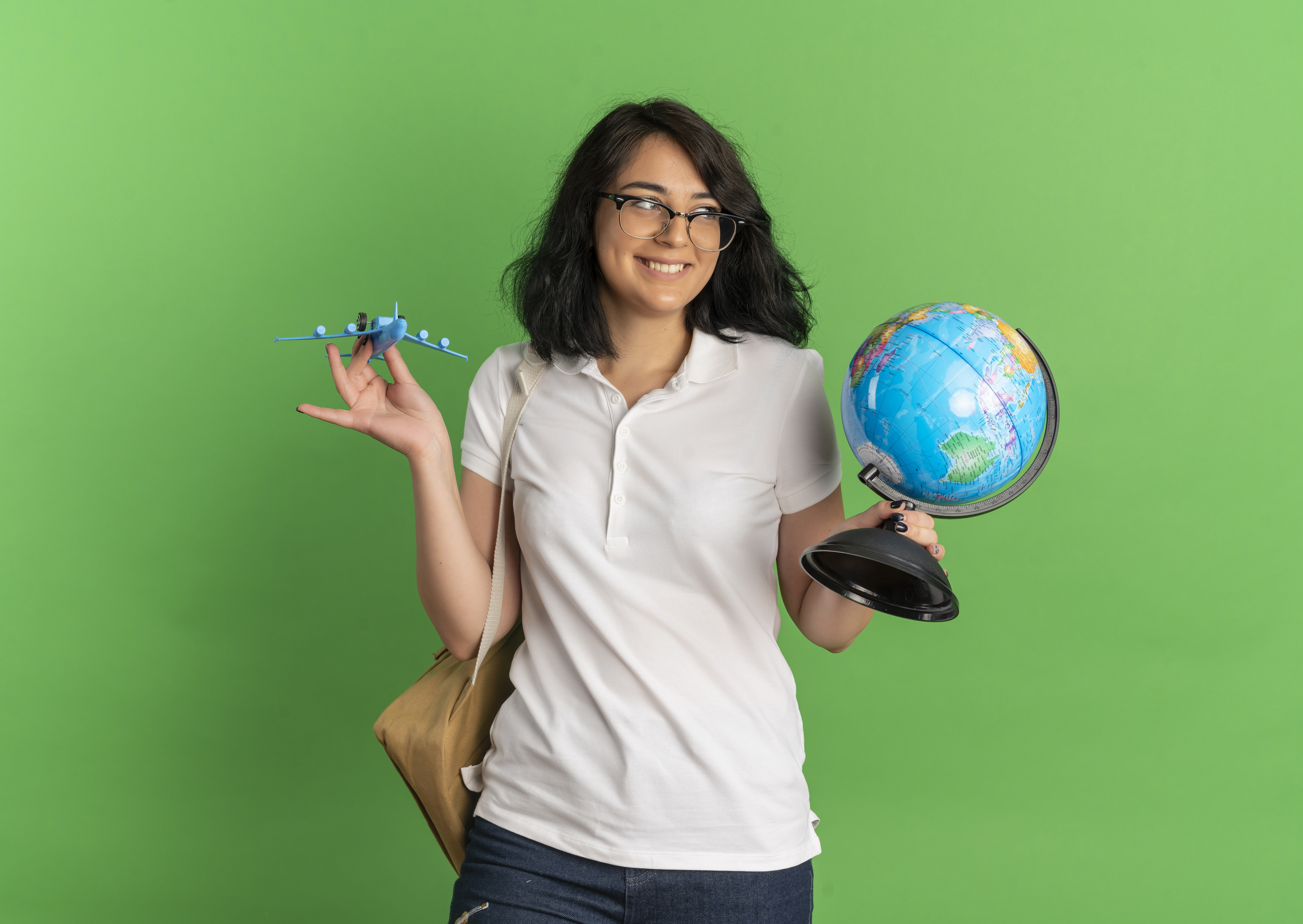 young smiling pretty caucasian schoolgirl wearing glasses and back bag holds toy plane and globe isolated on green background with copy space